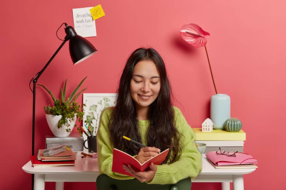 A person in a green sweater sits at a desk, writing in a red notebook, surrounded by plants, stationery, and decorative items against a pink wall.