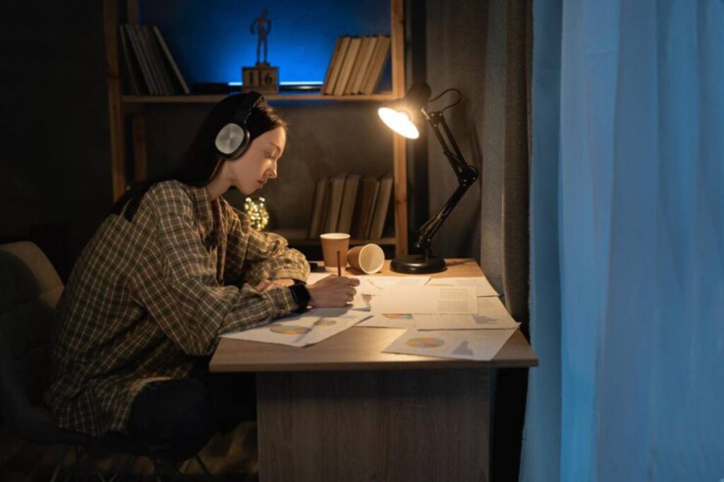 A person wearing headphones is seated at a desk covered with papers and a coffee cup, illuminated by a nearby desk lamp.