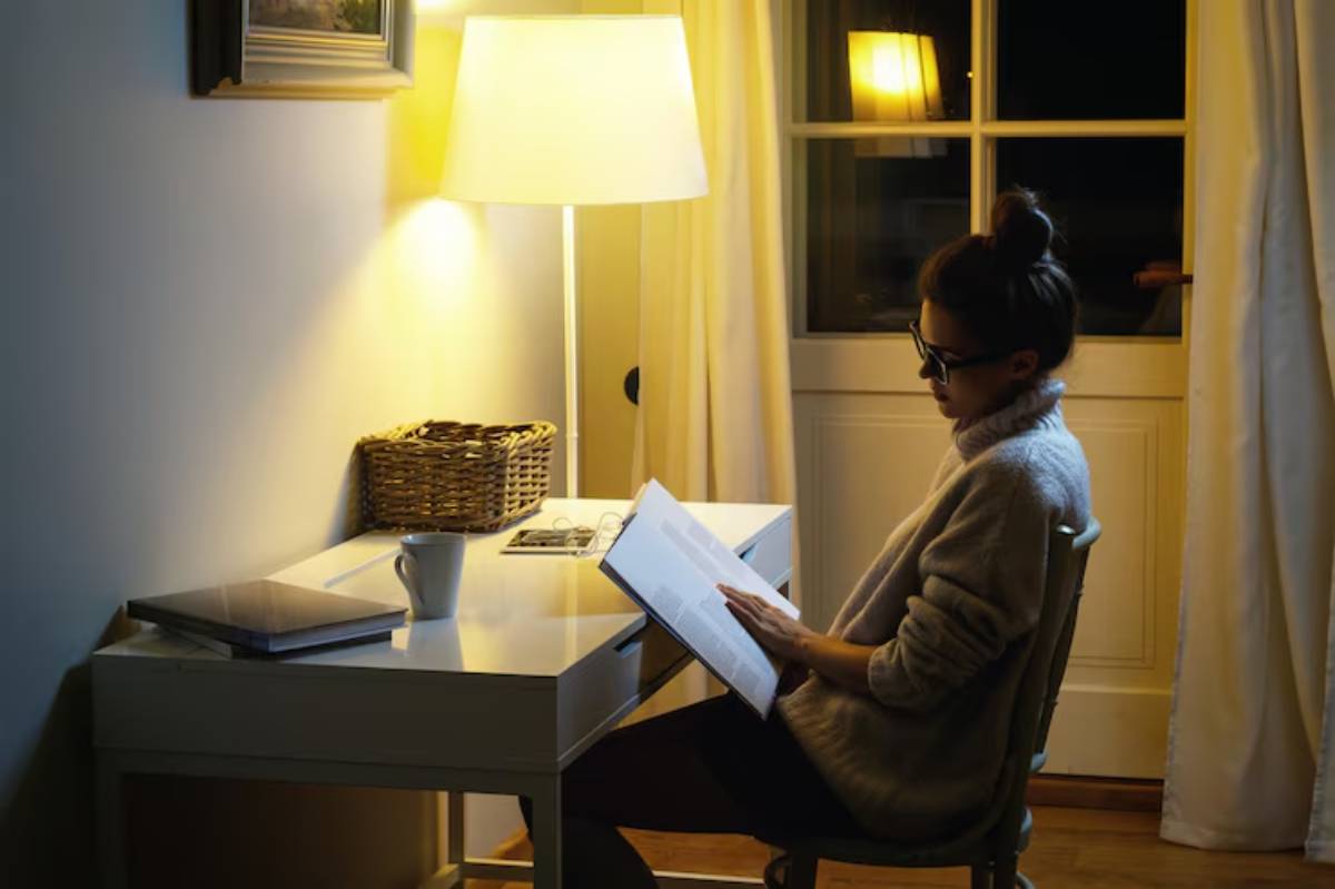 A person sits at a desk in a cozy room, reading a book under soft lamp light, with a cup and a laptop nearby.