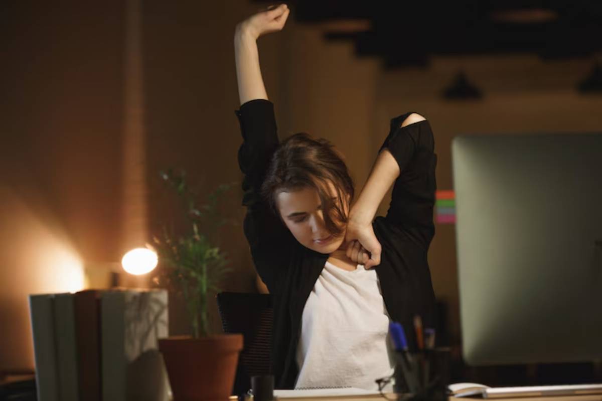 A person stretches their arms while sitting at a desk in a dimly lit room, surrounded by plants.