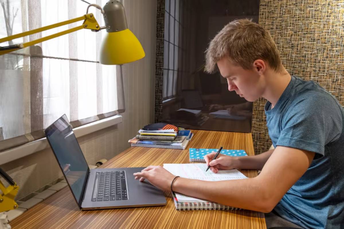 A person sits at a wooden desk, working on a laptop and taking notes in a notebook, surrounded by books and a yellow lamp.