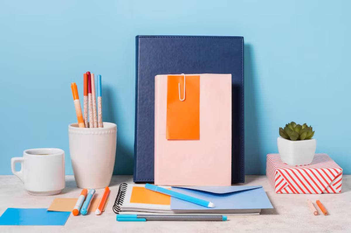 A bright room featuring a navy binder, a pink notebook with an orange tag, pens, sticky notes, a white mug, and a small potted plant.
