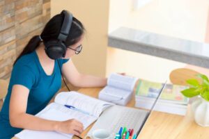 A student wearing headphones studies at a wooden desk, surrounded by books, a laptop, and stationery, focused on their work.
