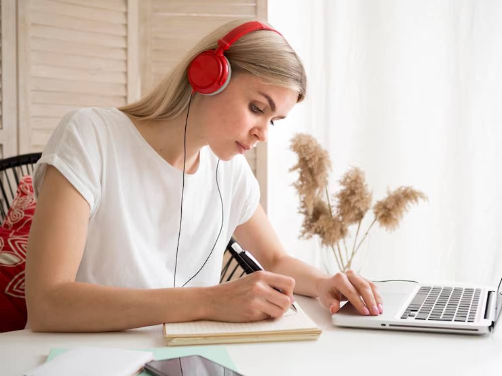 A person in a white shirt wearing red headphones writes in a notebook while using a laptop.