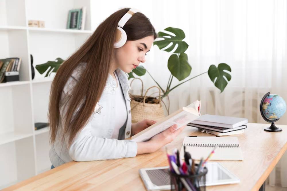 A person wearing headphones reads a book at a wooden desk, surrounded by notebooks, a globe, and a potted plant in a bright room.