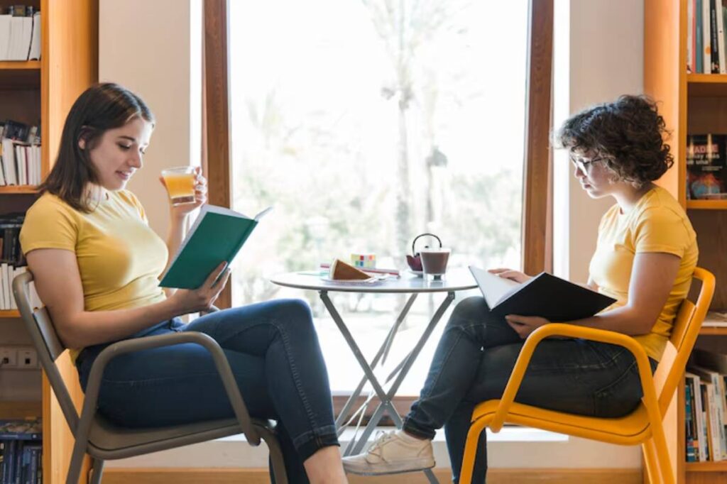 Two women in yellow shirts sit in a cozy cafe, engaged in reading and enjoying drinks at a small table by a window.