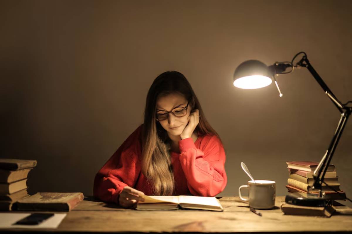 A person in a cozy red sweater sits at a wooden table, studying under the warm glow of a lamp surrounded by books and a mug.