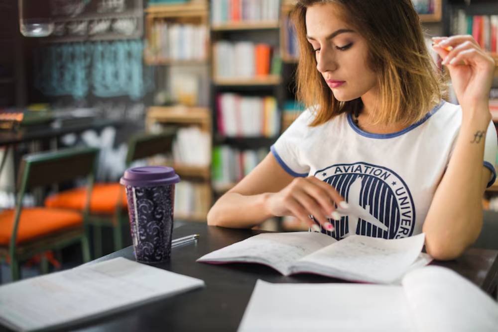 A student in an aviation-themed shirt studies at a café table, surrounded by books and holding a coffee cup.