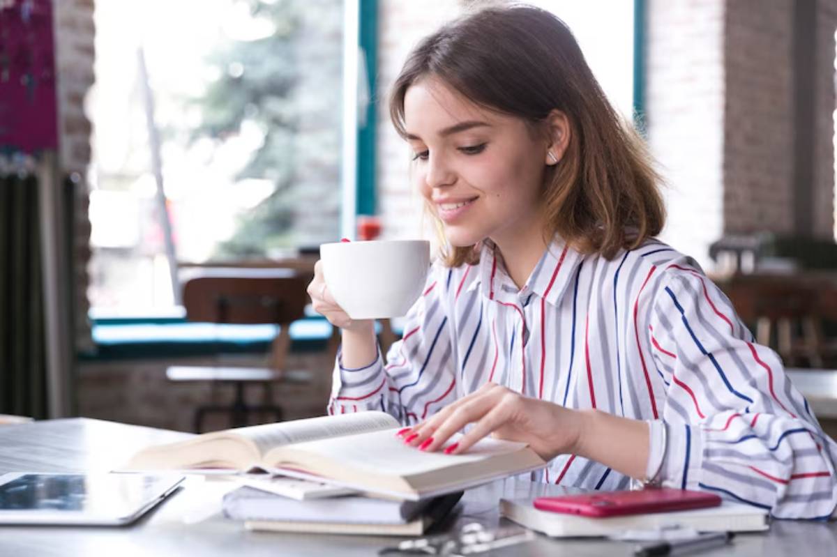 A person in a striped shirt sits at a table, enjoying a cup of coffee while reading a book in a cozy café setting.