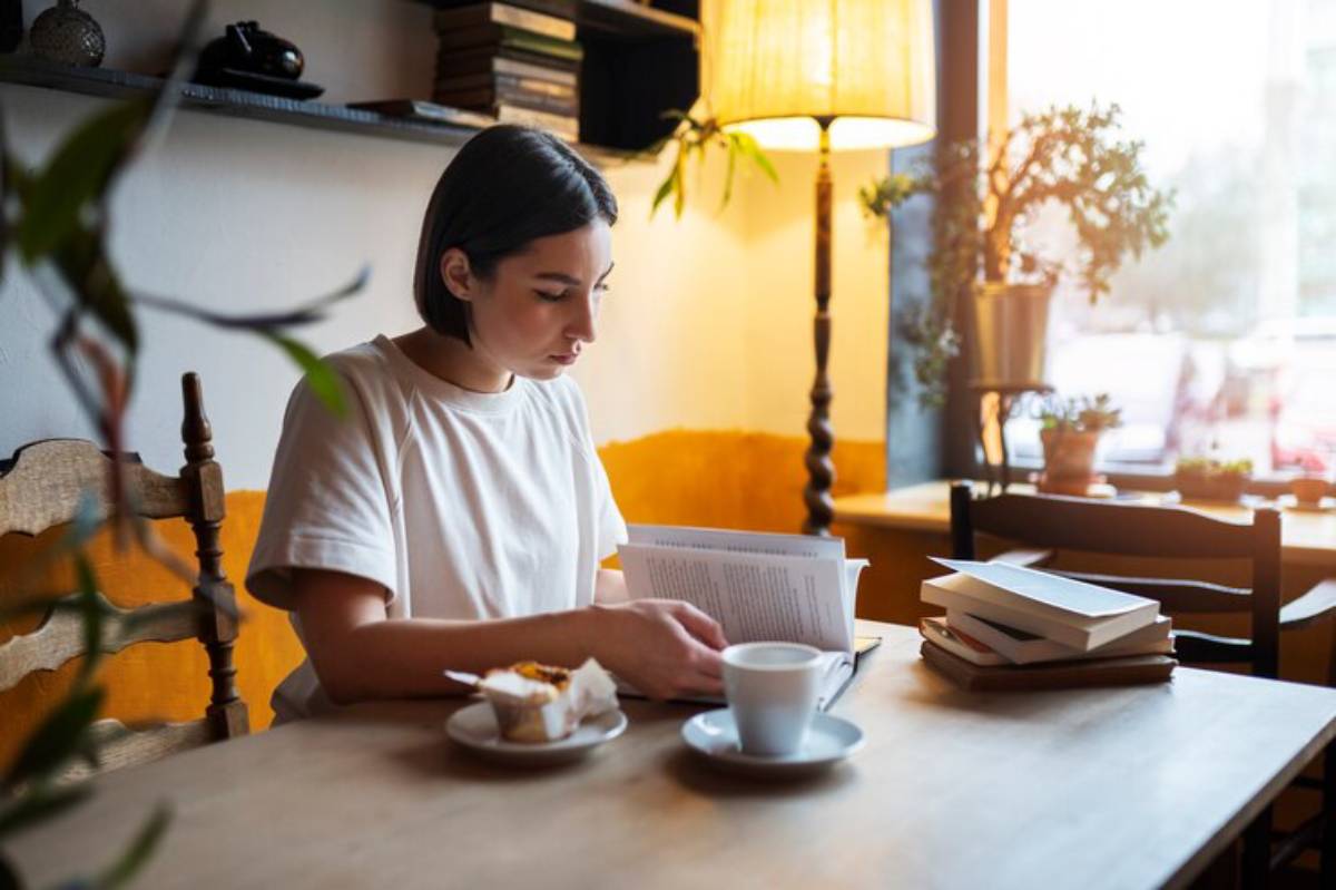 A person enjoys a cozy café moment, reading a book with a cup of coffee and pastry on the table, surrounded by warm light and plants.