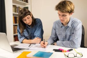 Two students study at a table, surrounded by books, with a laptop, notebooks, and colorful highlighters in front of them.