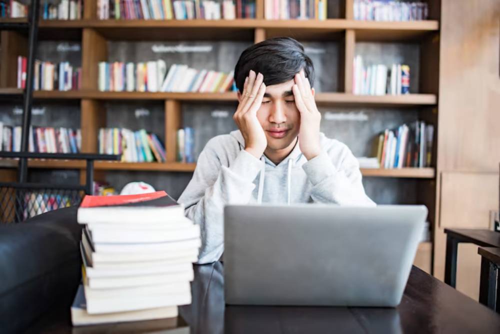 A young person in a gray hoodie sits at a desk with a laptop and a stack of books, showing signs of frustration in a cozy library-like setting.
