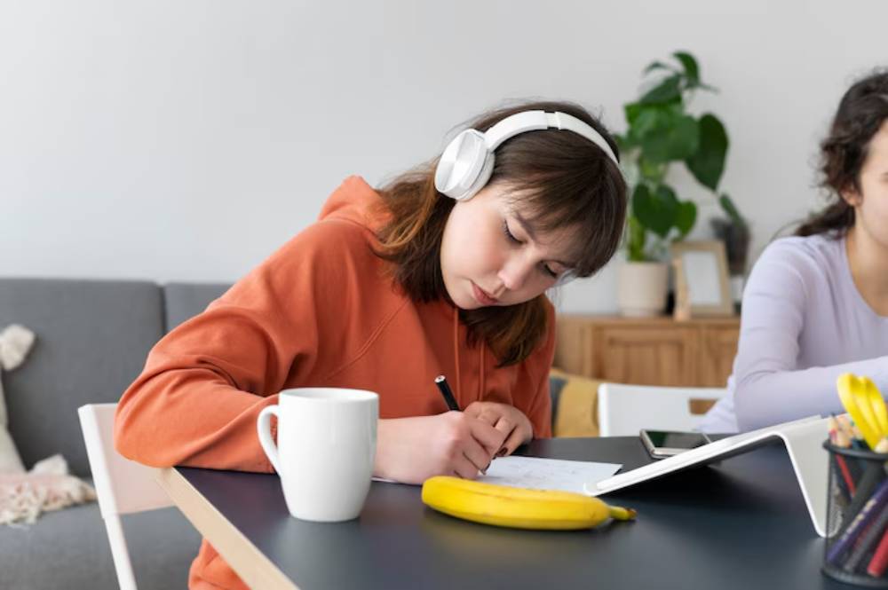 A person in an orange hoodie writes notes with headphones on, a cup and a banana on the table, while another person works nearby.