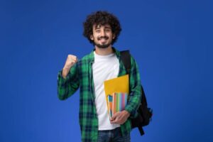 A young person with curly hair, wearing a green plaid shirt, holds colorful folders, showing an enthusiastic fist pump against a blue background.