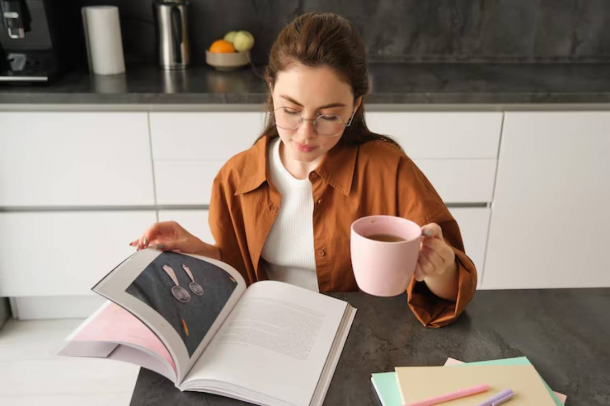 A person sits at a kitchen table, holding a pink cup of coffee while reading an open book.
