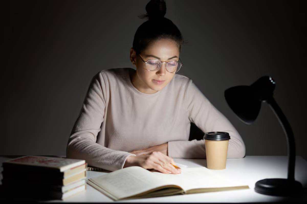 A person studies at a dimly lit table with an open book, coffee cup, and stack of books, illuminated by a desk lamp.