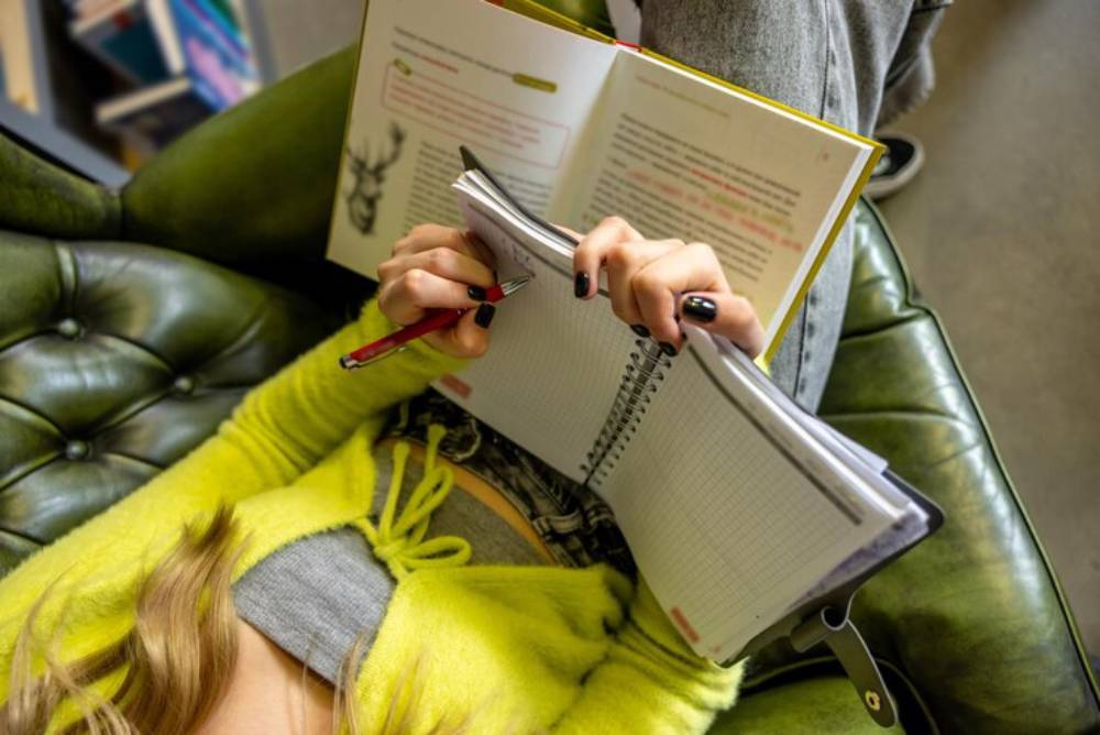 A person in a neon green sweater relaxes on a green chair, jotting notes in a notebook while reading an open book.