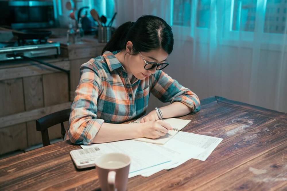 A person in a plaid shirt writes in a notebook at a wooden table, surrounded by papers and a cup, in a cozy kitchen setting.