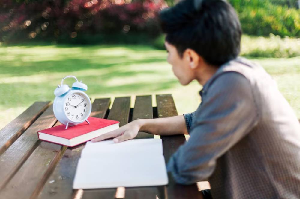 A young person sits at a wooden table outdoors, with a blue alarm clock and a red book next to a blank notebook on the table.