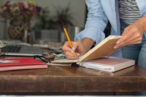 A person writes in a notebook with a pencil while surrounded by stationery and a laptop on a wooden desk.