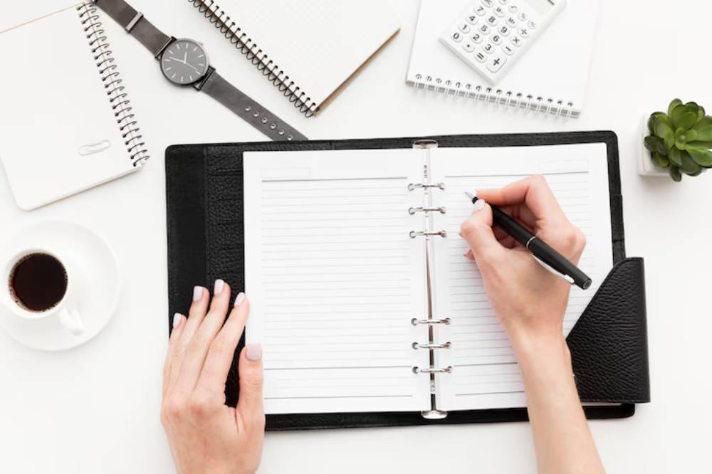 A hand writes in a planner on a desk with a coffee cup, notebooks, a calculator, a clock, and a small potted plant.