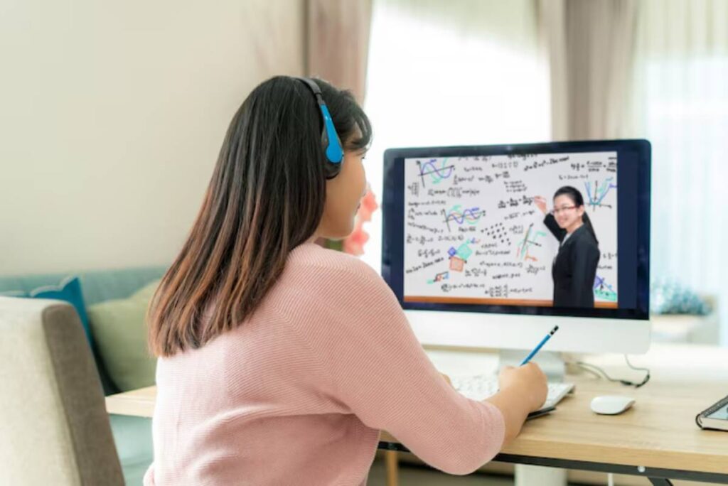 A student in a pink sweater attentively participates in an online class, taking notes while viewing mathematical diagrams on a monitor.
