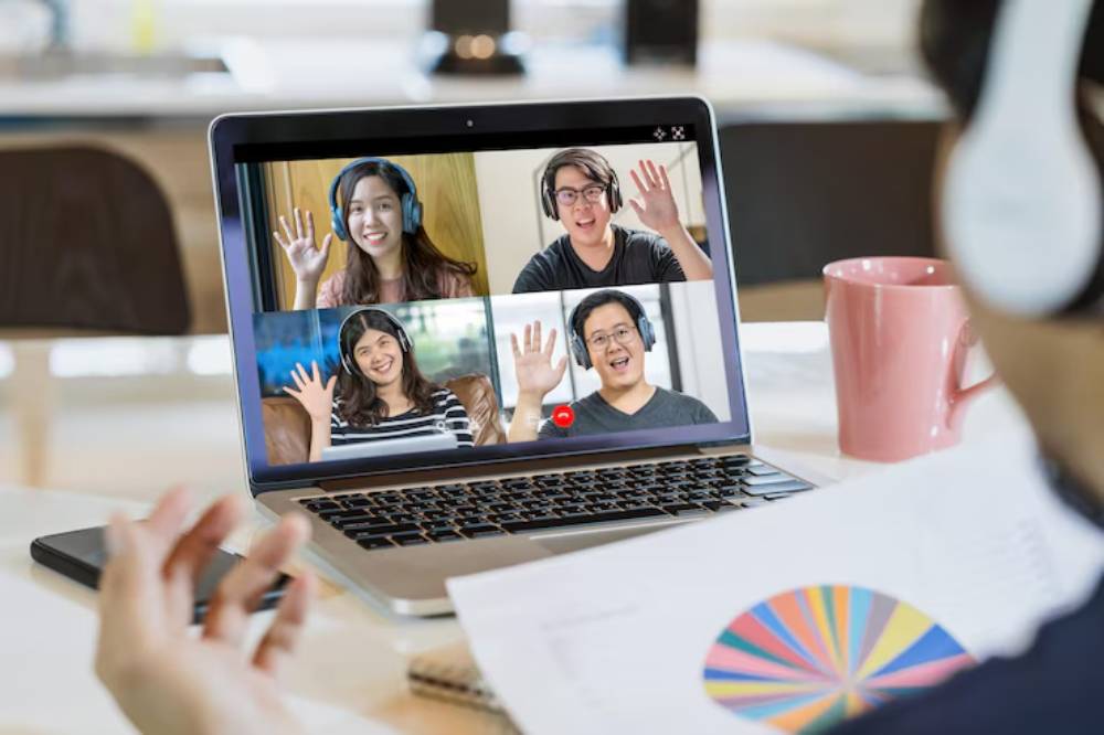 A person participates in a virtual meeting on a laptop, displaying multiple attendees, each wearing headphones and waving. A pink mug and papers are nearby.