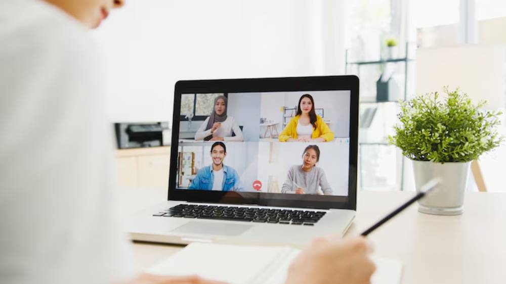 A person participates in a video call on a laptop, with multiple participants displayed on the screen and a small plant nearby.