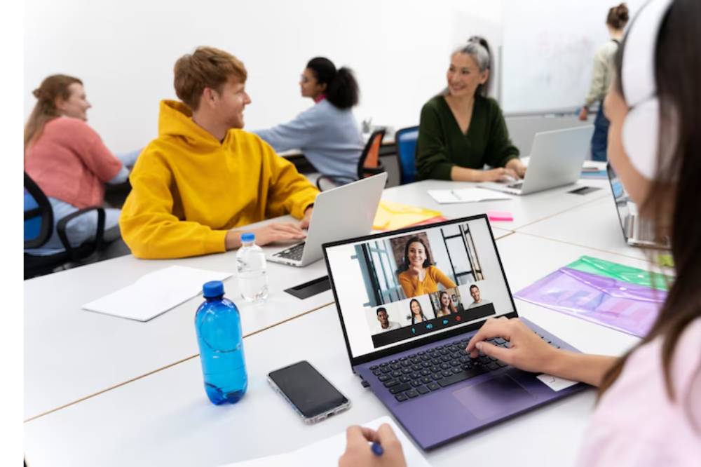 A group of students engaged in a virtual class, using laptops and notebooks in a modern classroom setting. Water bottles are present.