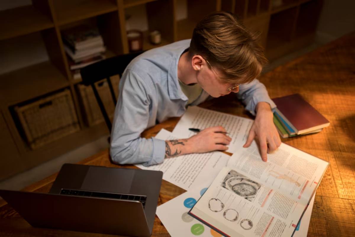 A person studies at a wooden table, surrounded by open books, notes, and a laptop, with a focus on detailed diagrams and text.