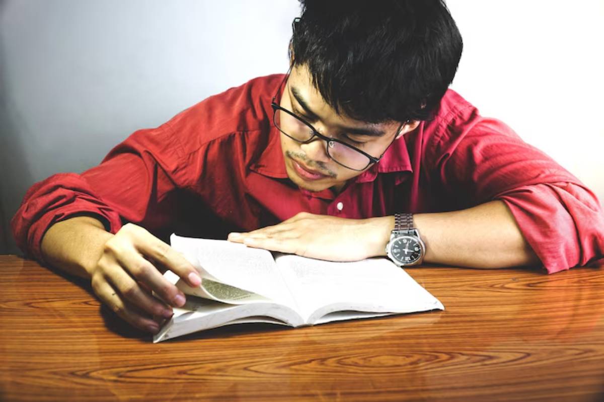 A person in a red shirt reading a book, sitting at a wooden table, with a watch visible on their wrist.