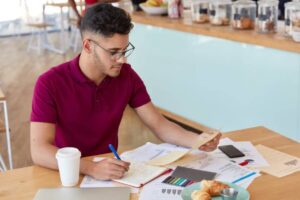 A person in a red polo shirt takes notes and reviews papers at a wooden table with coffee, pastries, and a laptop.