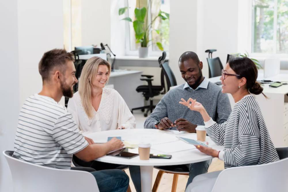 A group of four people engages in a discussion around a round table in a bright, modern office space with greenery.