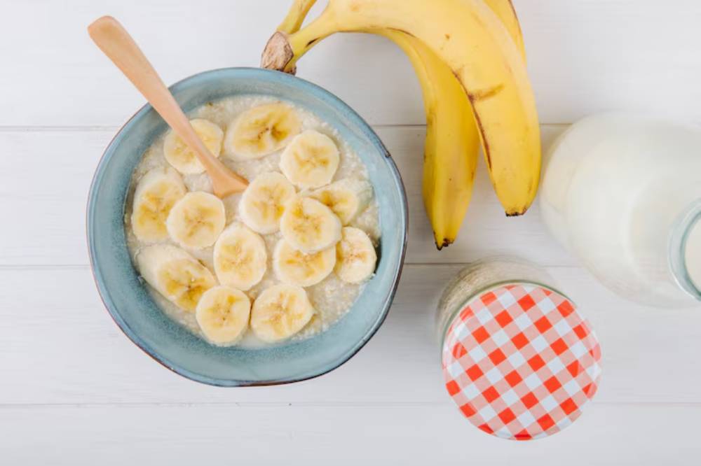 A bowl of oatmeal topped with banana slices and a wooden spoon, next to fresh bananas and a jar with a checkered lid.