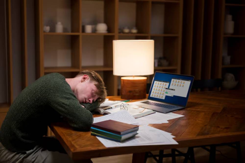 A person rests their head on a cluttered desk, surrounded by books and papers, with a laptop displaying folders and a lamp illuminating the scene.