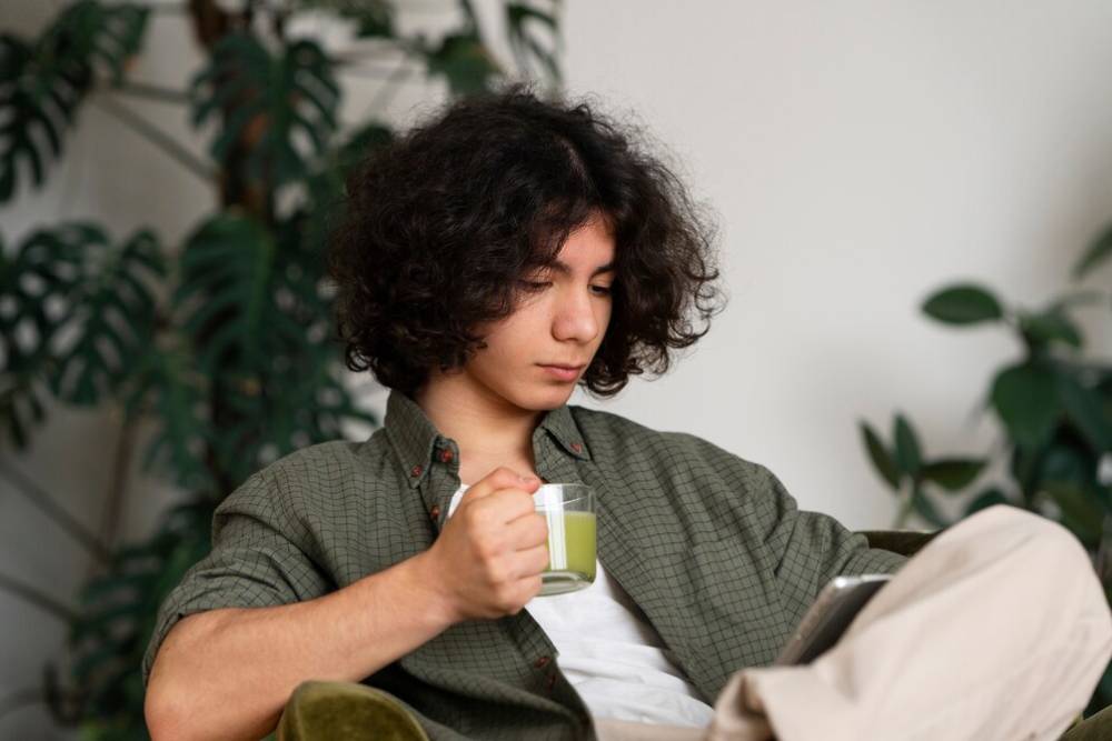 A person with curly hair sits comfortably in a green chair, holding a glass of green tea, surrounded by indoor plants.